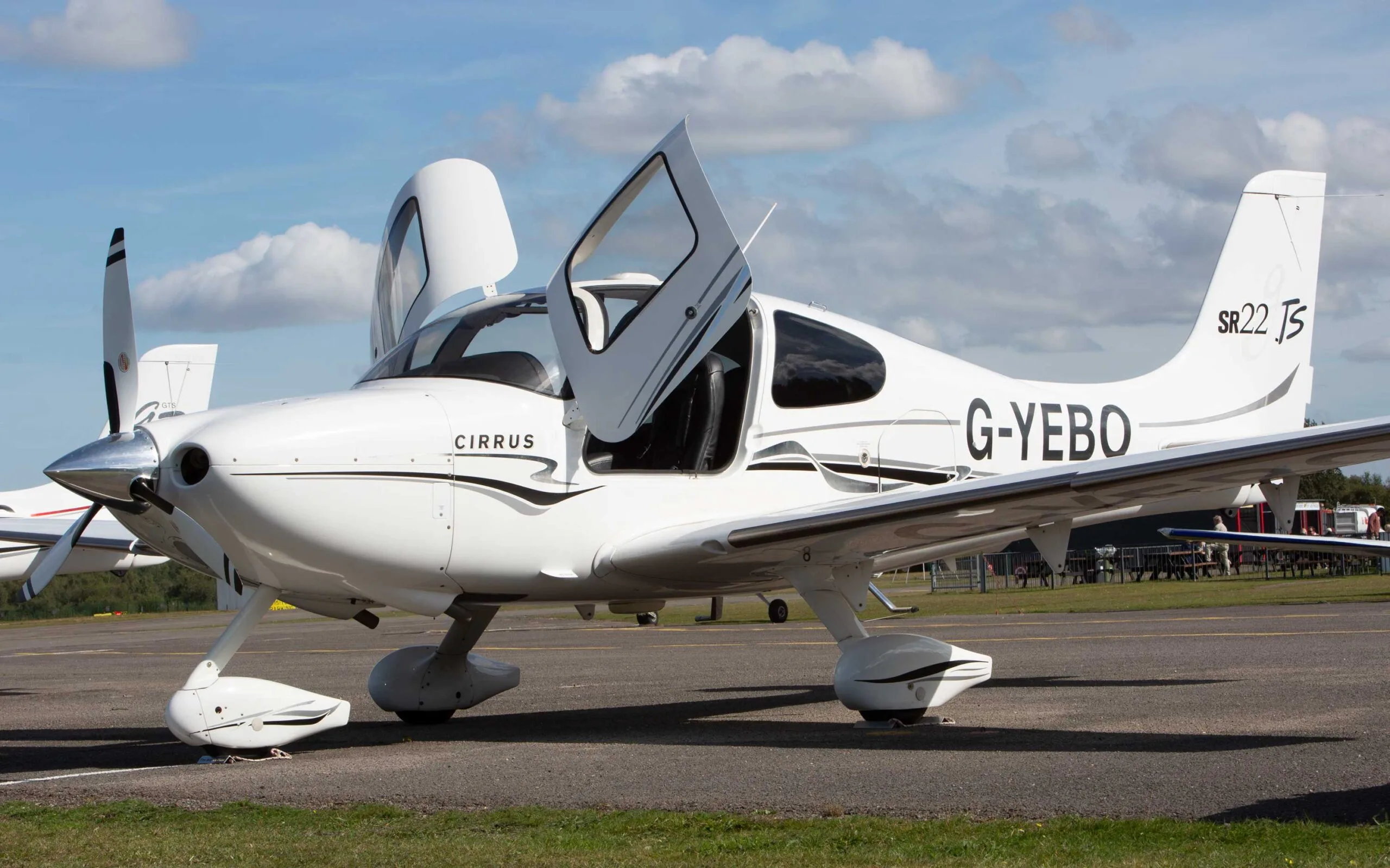 A white Cirrus SR22 TSi aircraft with black and grey stripes, registration G-YEBO, parked on an airfield with the right gull-wing door open.