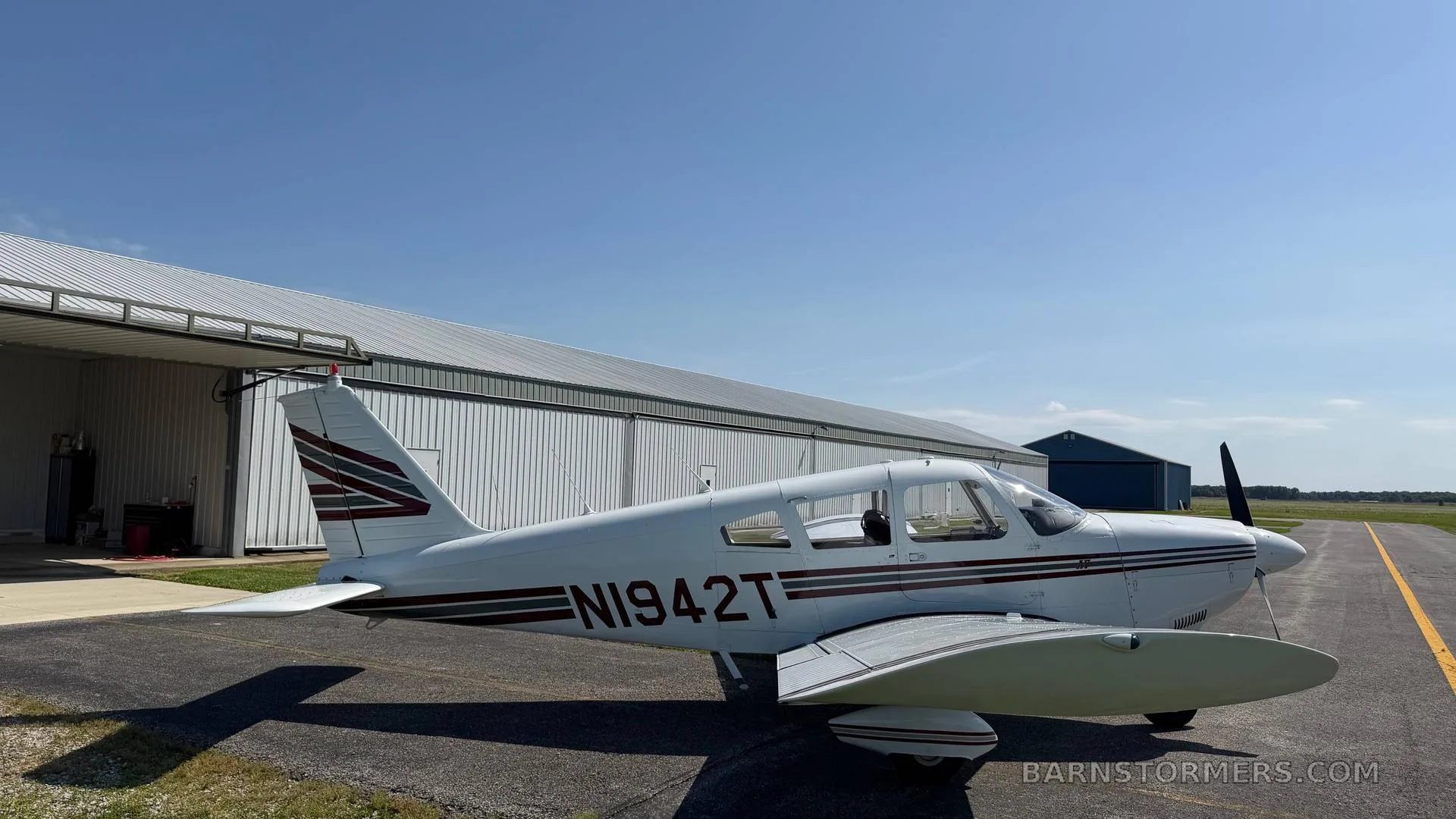 A white single-engine propeller aircraft (N1942T) with red and grey stripes parked on an asphalt ramp in front of a large metal hangar.