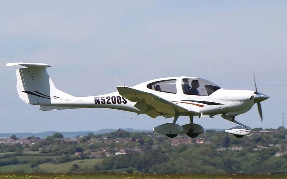 A white Diamond DA40 aircraft with brown and black stripes flying low over a green landscape.