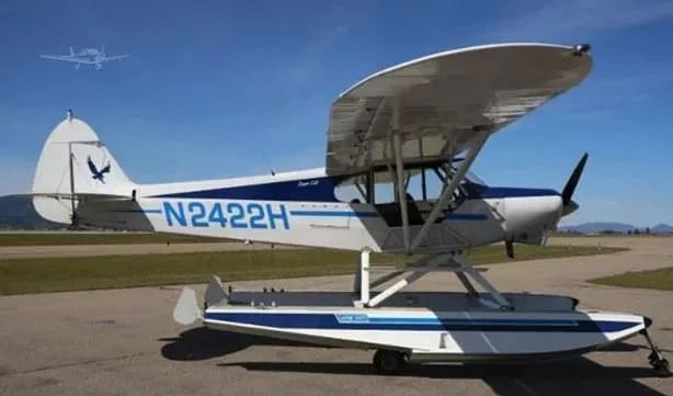 A white and blue floatplane with registration N2422H is parked on an airfield under a clear blue sky.