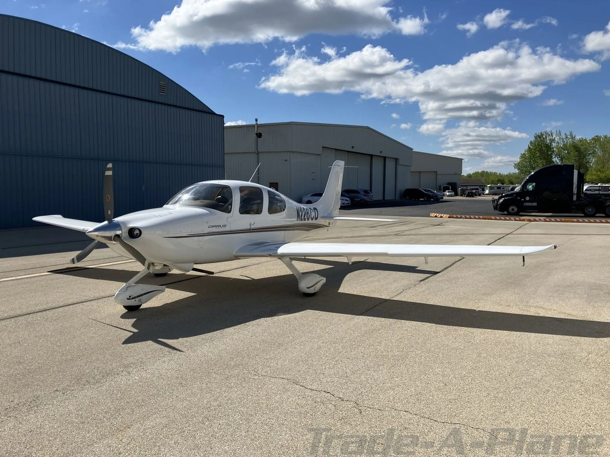 A white Cirrus SR20 aircraft with black and brown stripes, tail number N226CD, parked on an airfield tarmac next to a blue hangar.