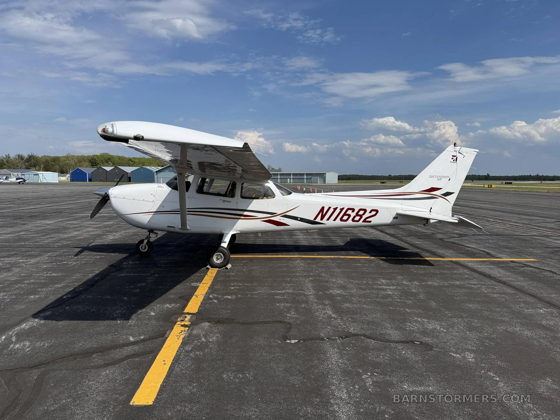 A white Cessna 172 aircraft with brown and black stripes, registration N11682, parked on an asphalt tarmac under a partly cloudy sky.