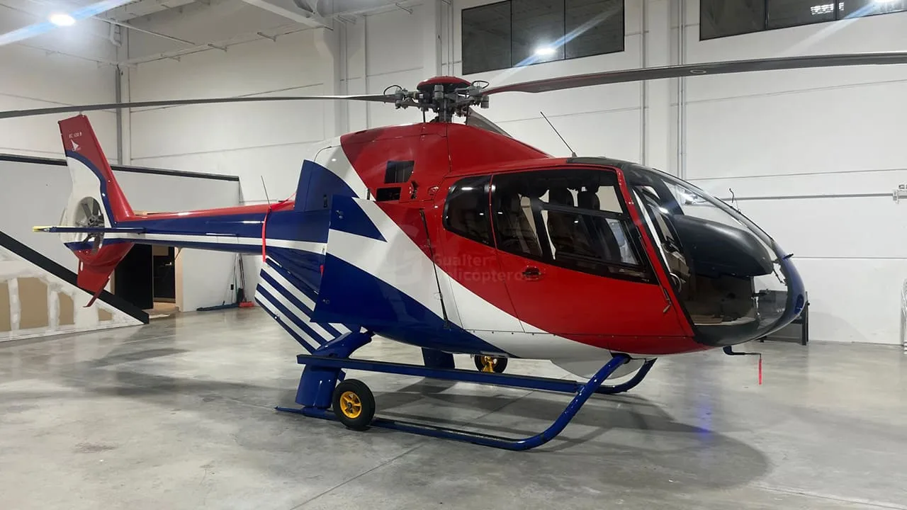 A red, white, and blue helicopter (likely an Airbus H125) parked inside a hangar.