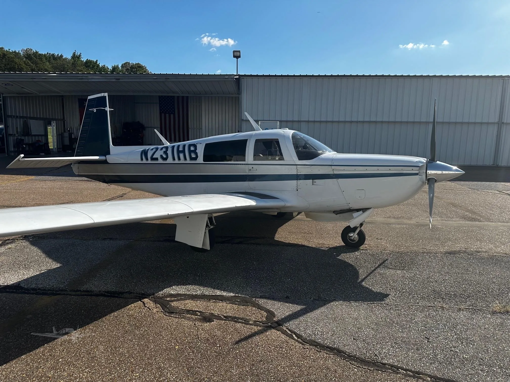 A white single-engine propeller aircraft with a dark blue stripe is parked on an asphalt apron next to a hangar.