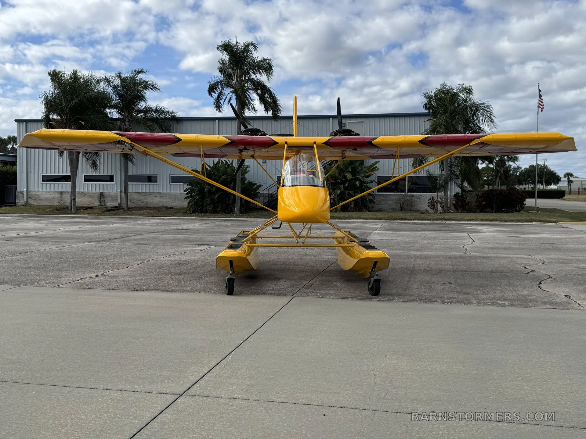 A yellow and red Air-Cam seaplane with floats, viewed from the front, parked on an asphalt ramp with buildings and palm trees in the background.
