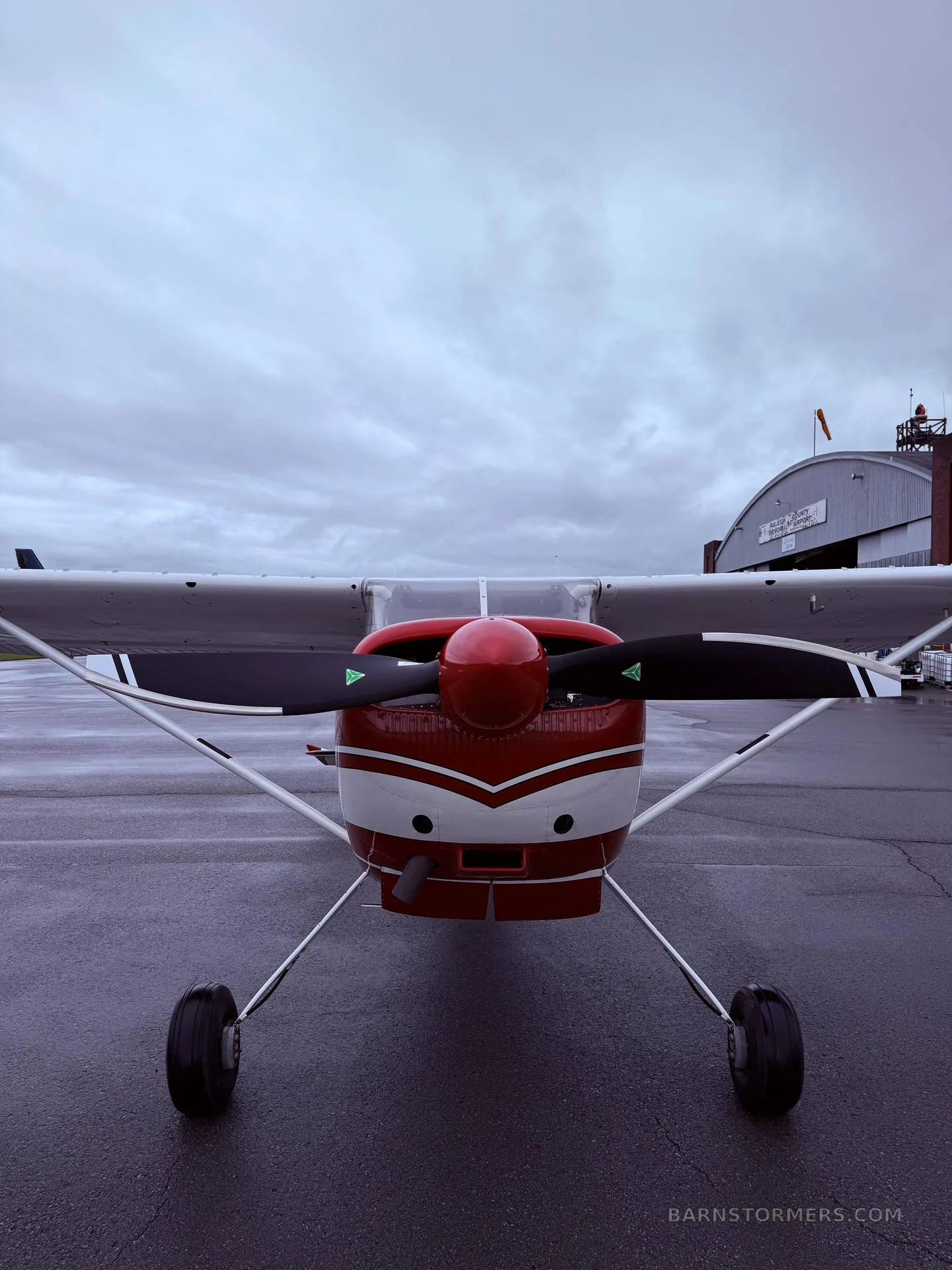 A red and white single-engine propeller aircraft is parked on a wet tarmac, viewed directly from the front.