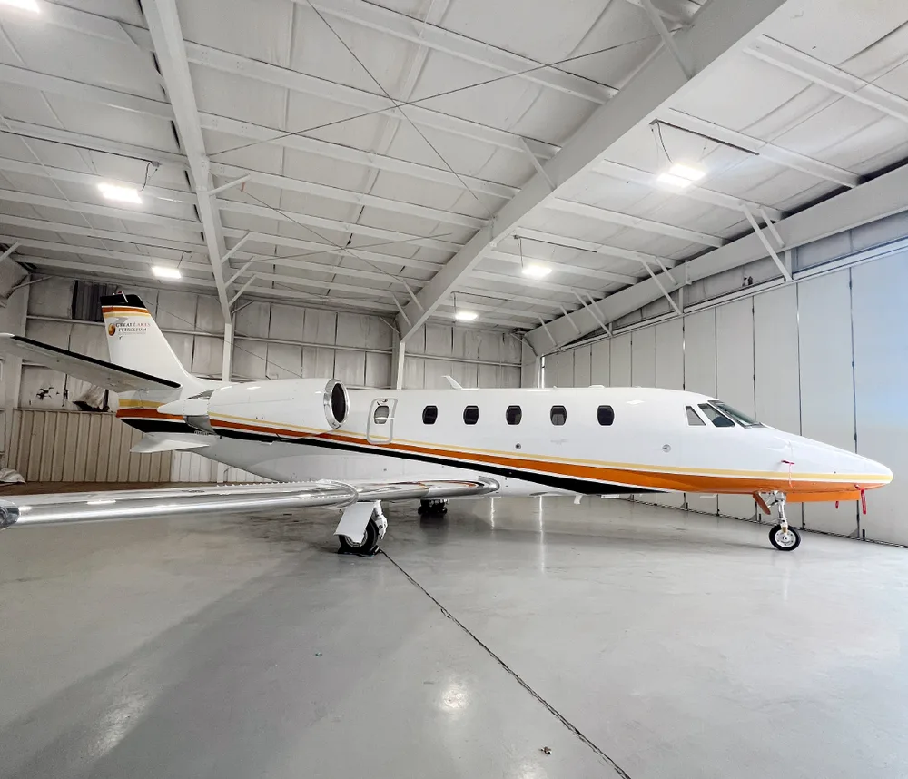 A white business jet with orange and black stripes, viewed from the front-right, inside a hangar.
