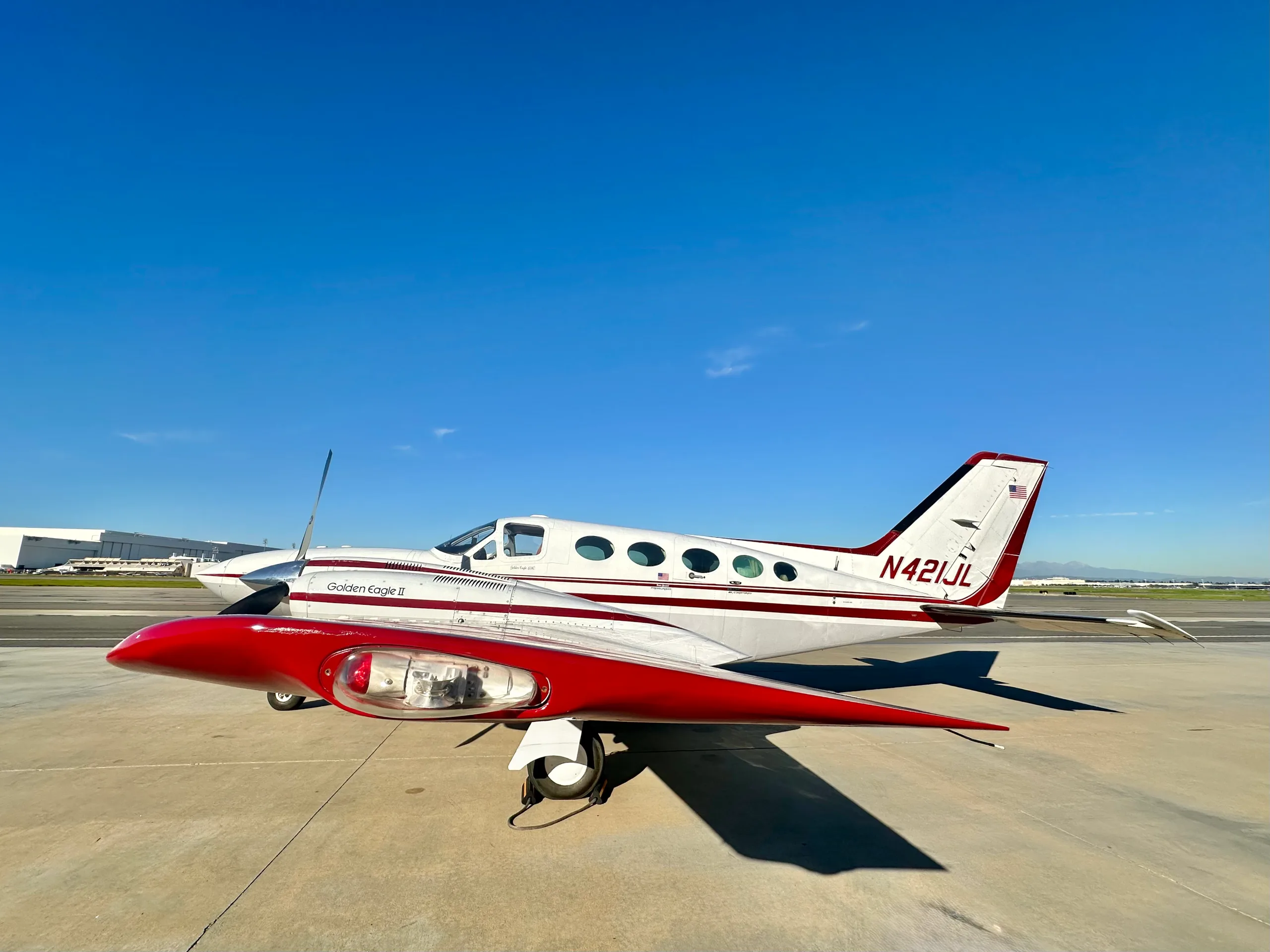 A white and maroon Cessna 421 Golden Eagle II, registration N421JL, with red wingtip tanks, parked on an airfield under a clear blue sky.
