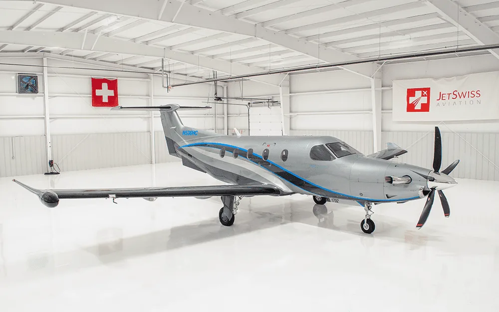 A silver Pilatus PC-12 turboprop aircraft with blue and grey stripes is parked inside a white hangar with Swiss flags and JetSwiss Aviation branding.