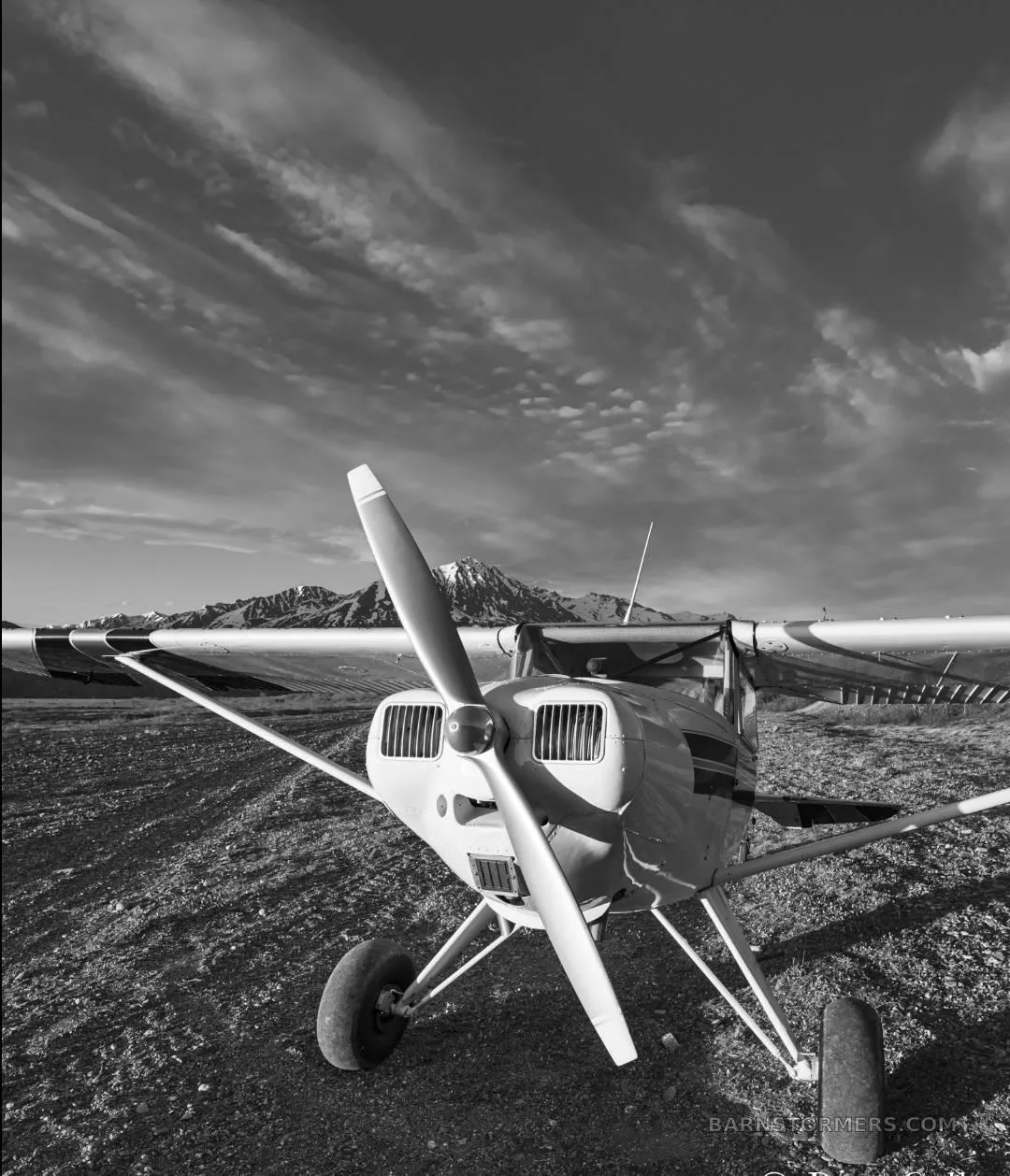 A black and white low-angle front view of a high-wing aircraft with large tires on a dirt field, with mountains under a cloudy sky.