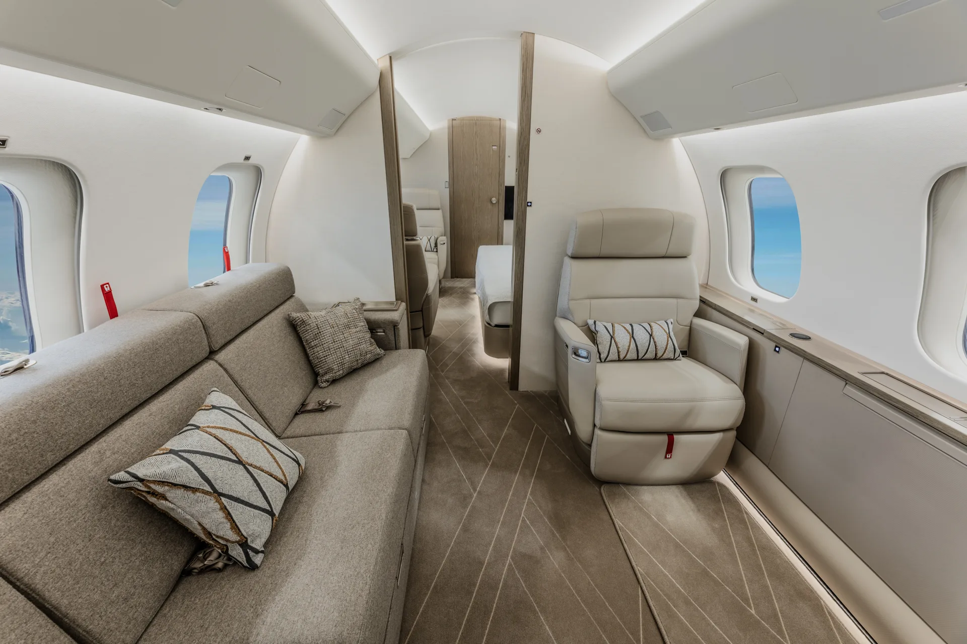 A view inside a private jet cabin showing a long grey sofa with pillows and a single cream leather seat.