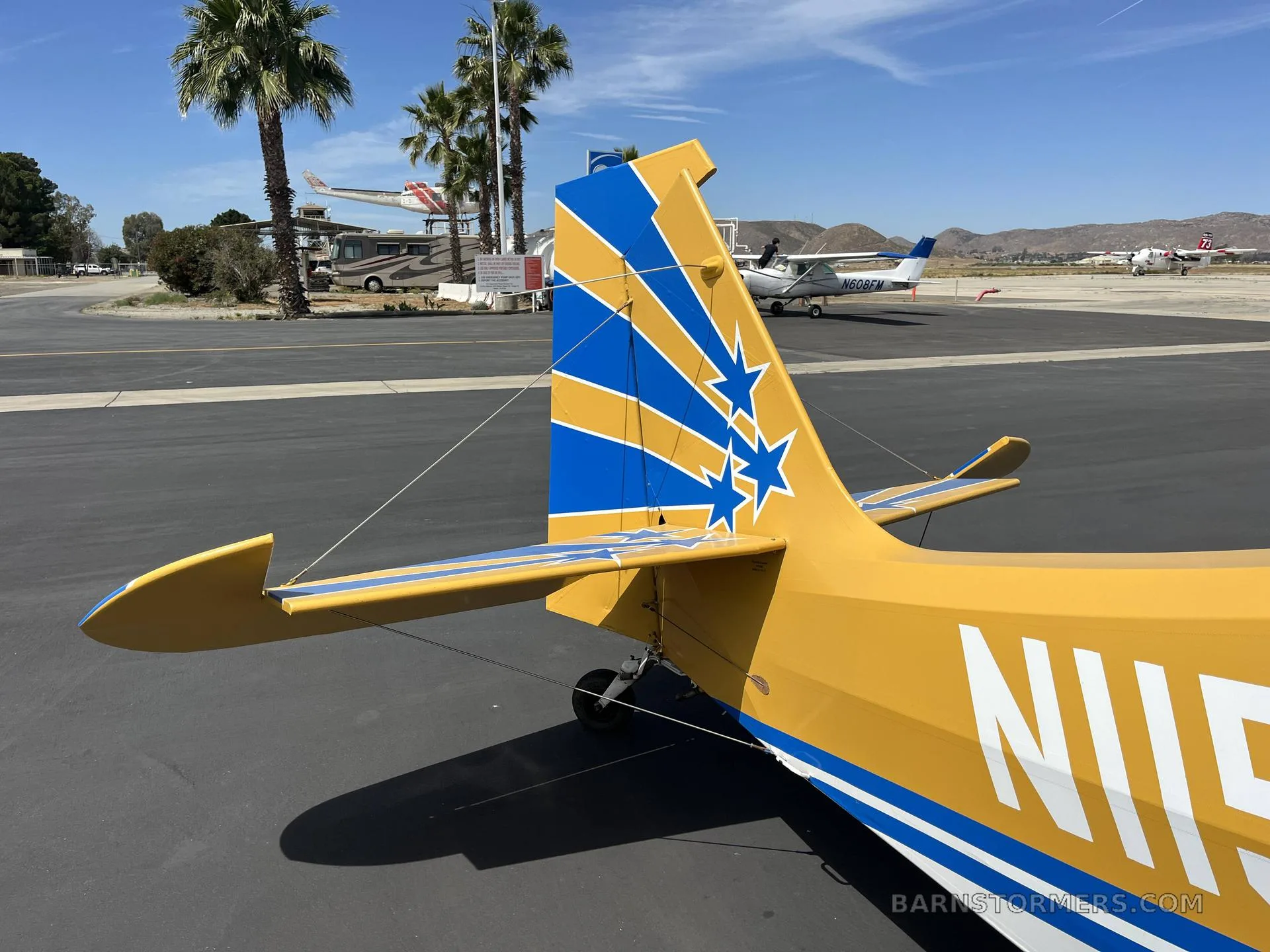 Rear view of a yellow aircraft's tail with blue stripes and stars, parked on an airport tarmac with other aircraft and palm trees in the background.