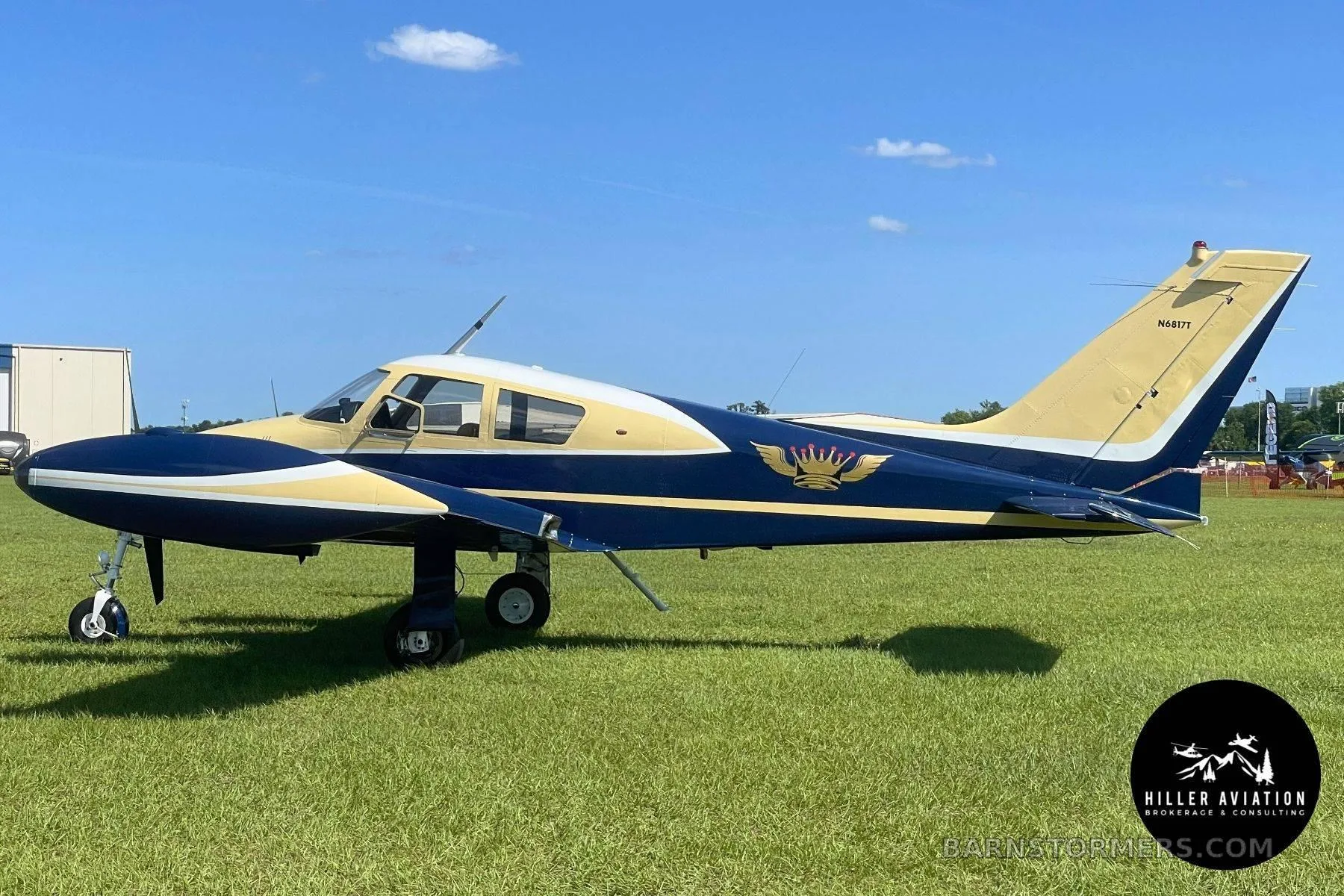 A blue and gold twin-engine aircraft with a crown logo on its side is parked on a grassy field under a blue sky.