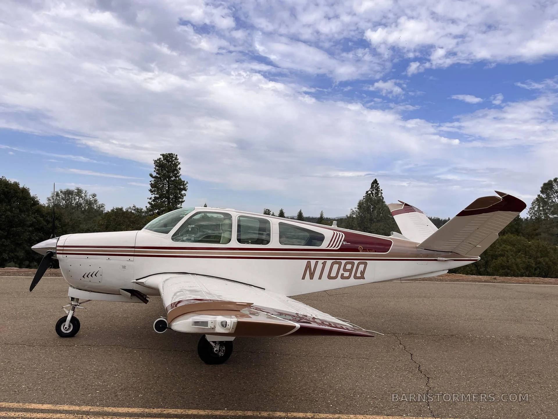 A white and brown Beechcraft Bonanza with registration N109Q, parked on an asphalt surface with trees and a cloudy sky in the background.