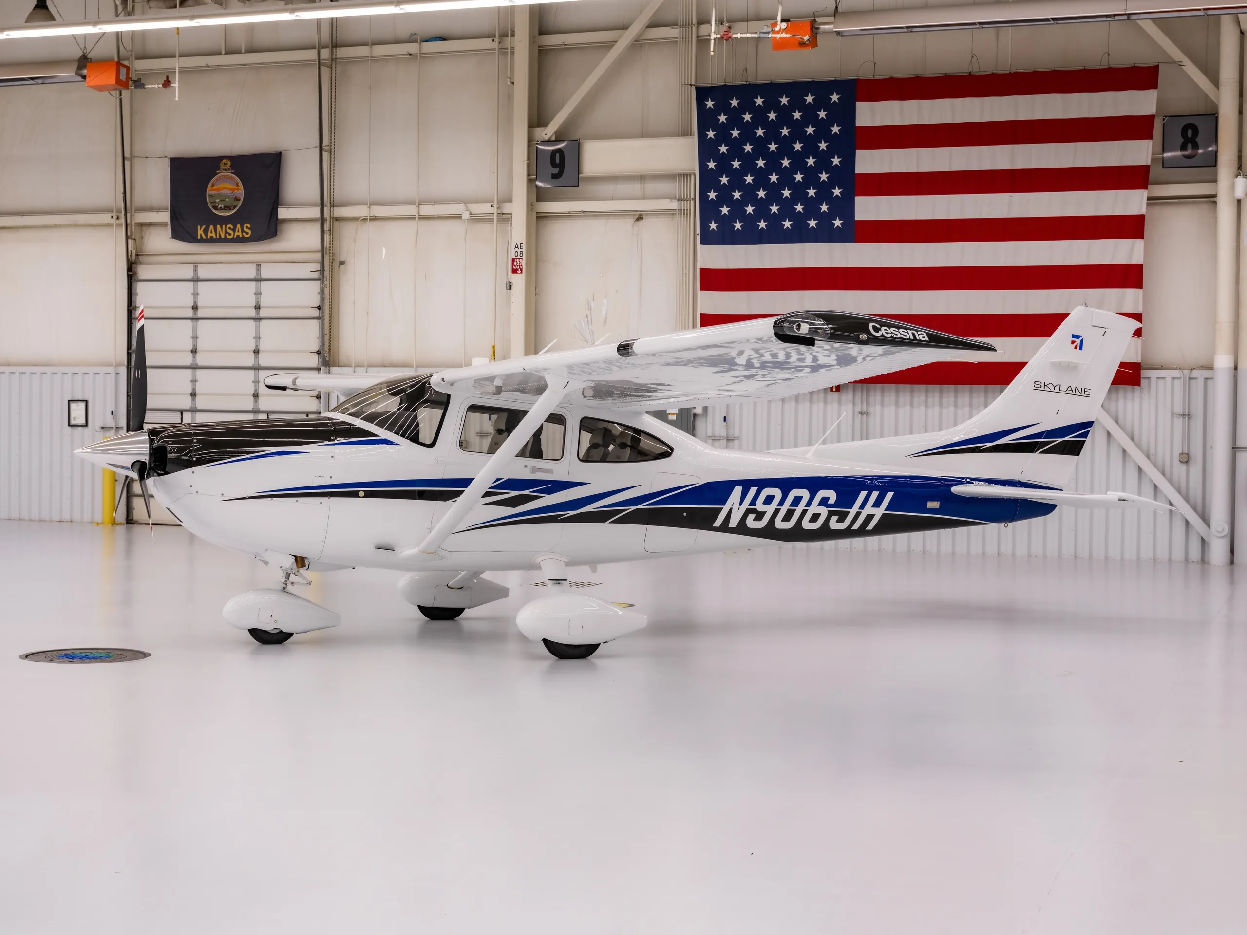 A white Cessna Skylane with blue and black stripes, registration N906JH, is parked inside a hangar with an American flag.