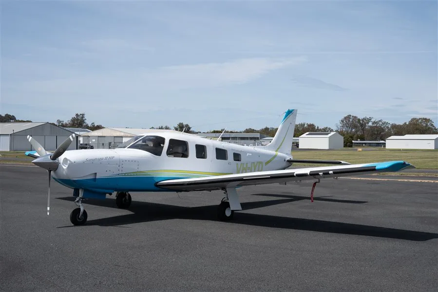 A white and blue Piper Seneca II HP aircraft (VH-IYD) parked on an asphalt tarmac with hangars in the background.