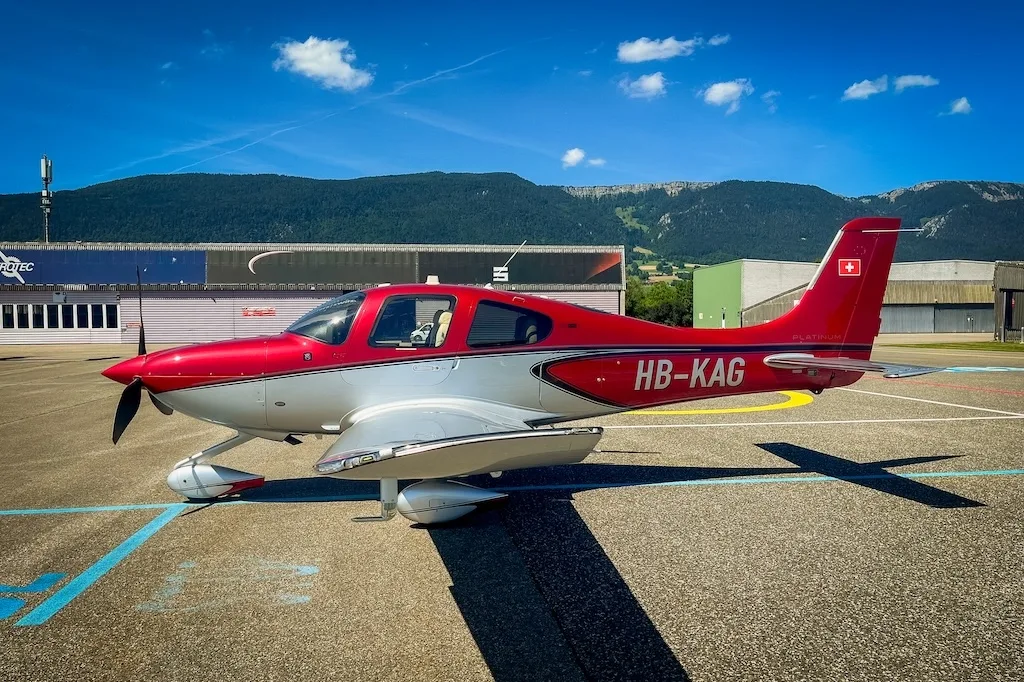 Red and silver Cirrus SR22 aircraft parked on an airfield, showing its side profile against a backdrop of mountains and buildings.