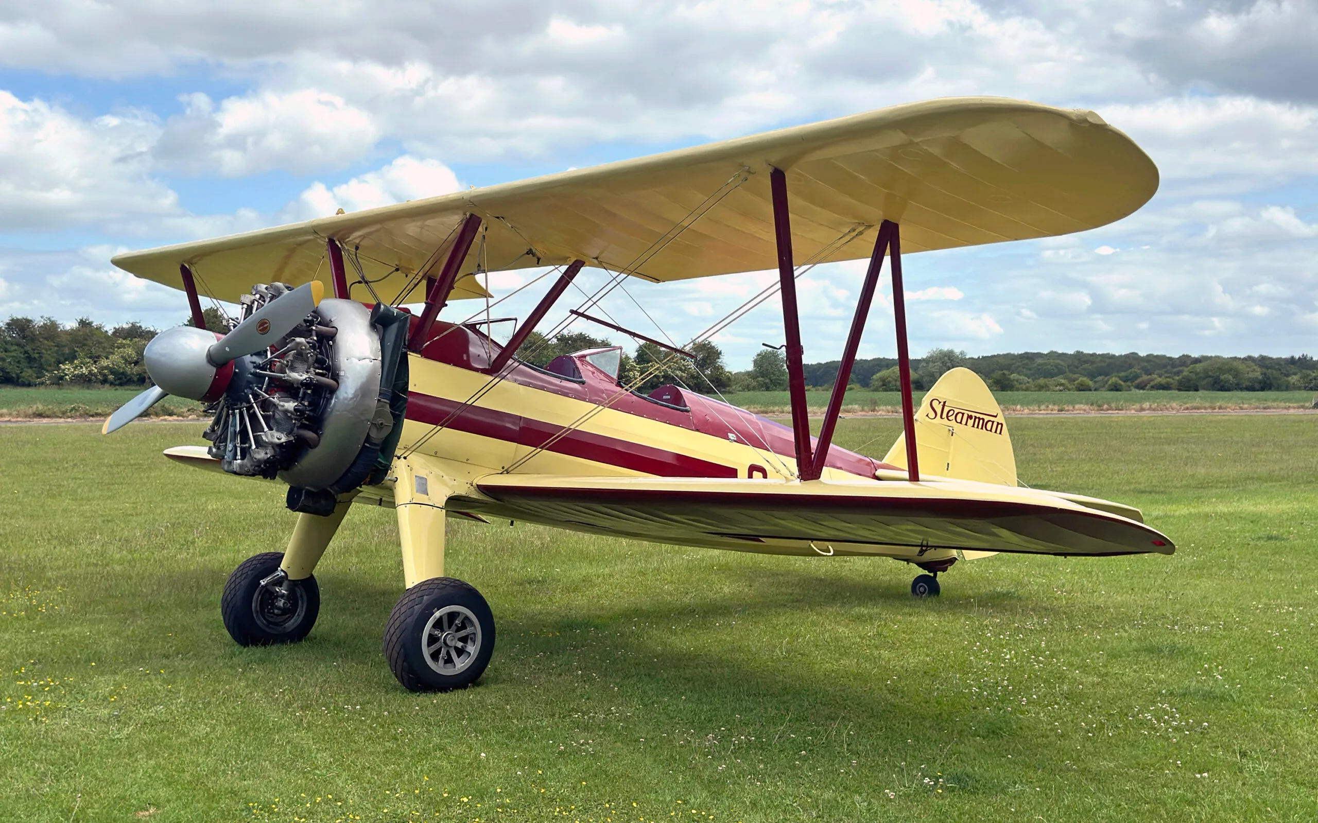 A yellow and red Stearman biplane, G-AROY, with its radial engine visible, parked on a green field.