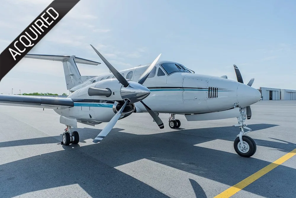 A white and blue twin-turboprop aircraft, likely a King Air, parked on an airport tarmac with an 'ACQUIRED' banner.