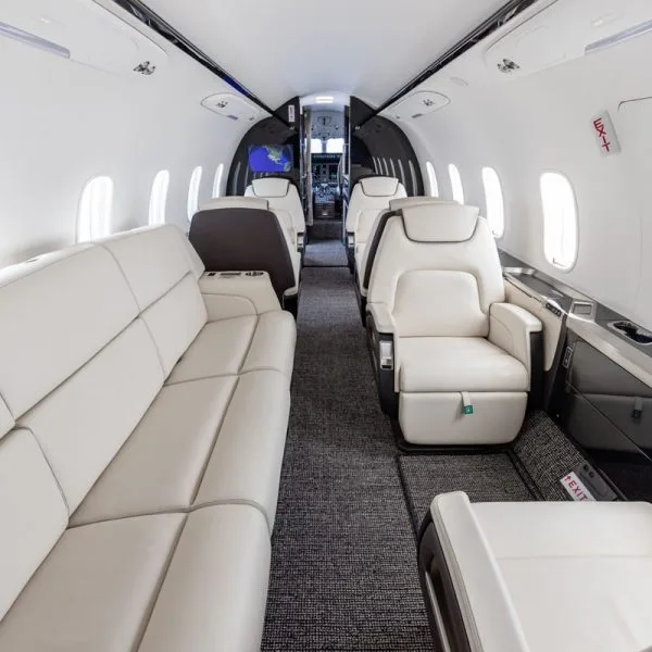 A wide interior view of a private jet cabin with a cream leather sofa and individual seats, looking towards the cockpit.