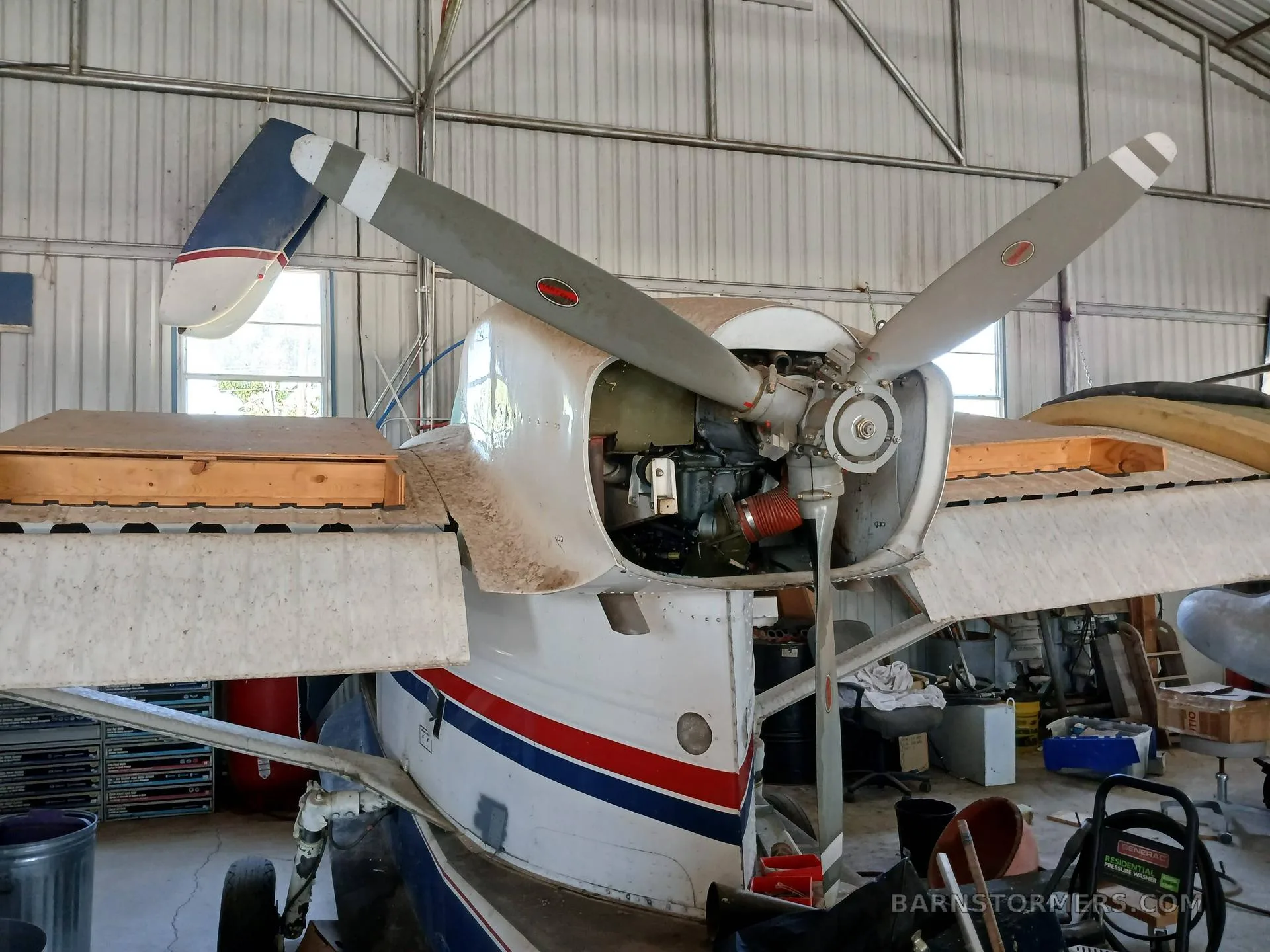 Front view of the engine and propeller of a white, red, and blue amphibious aircraft, with the cowling removed, inside a hangar.