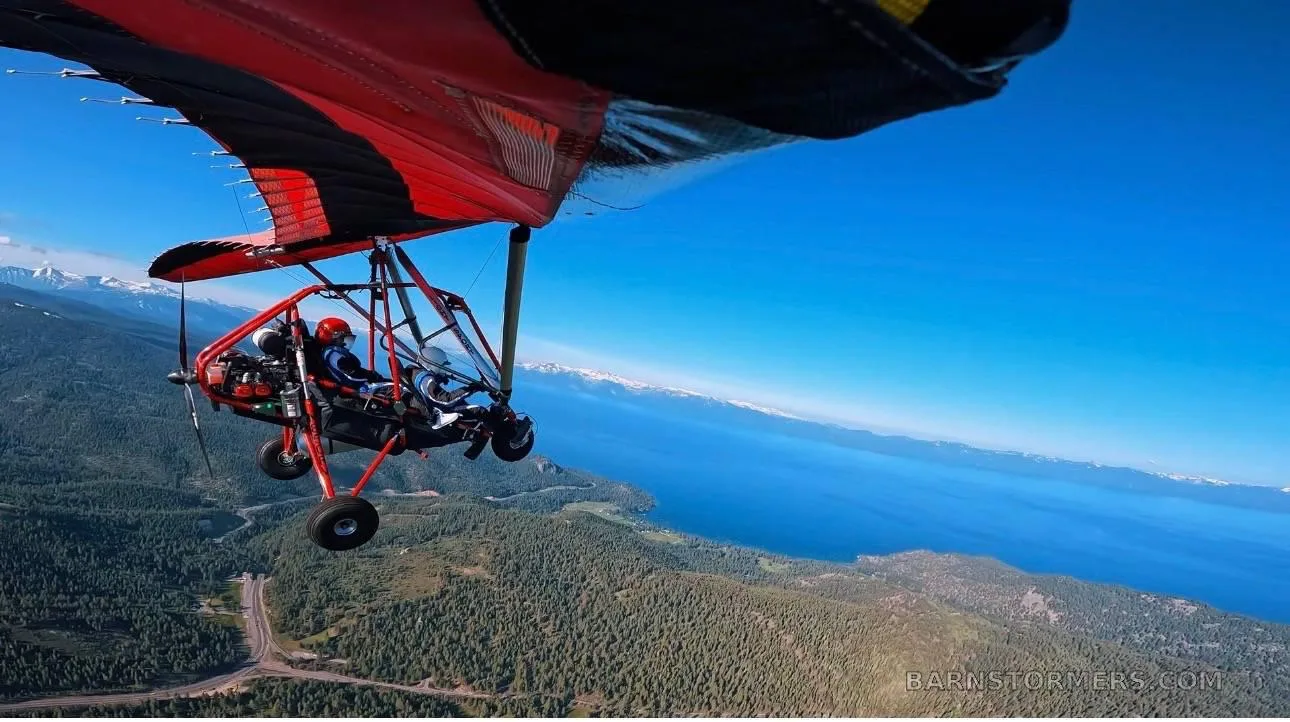 A red and black trike ultralight aircraft with two occupants flying over a forested landscape next to a large lake and distant snow-capped mountains.
