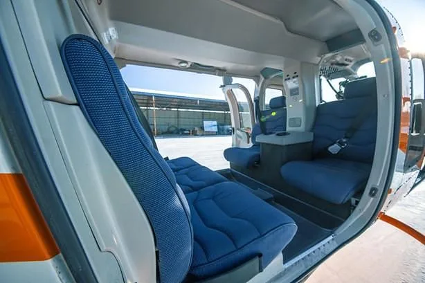 The interior cabin of a helicopter features blue fabric seats and an open door revealing a hangar.