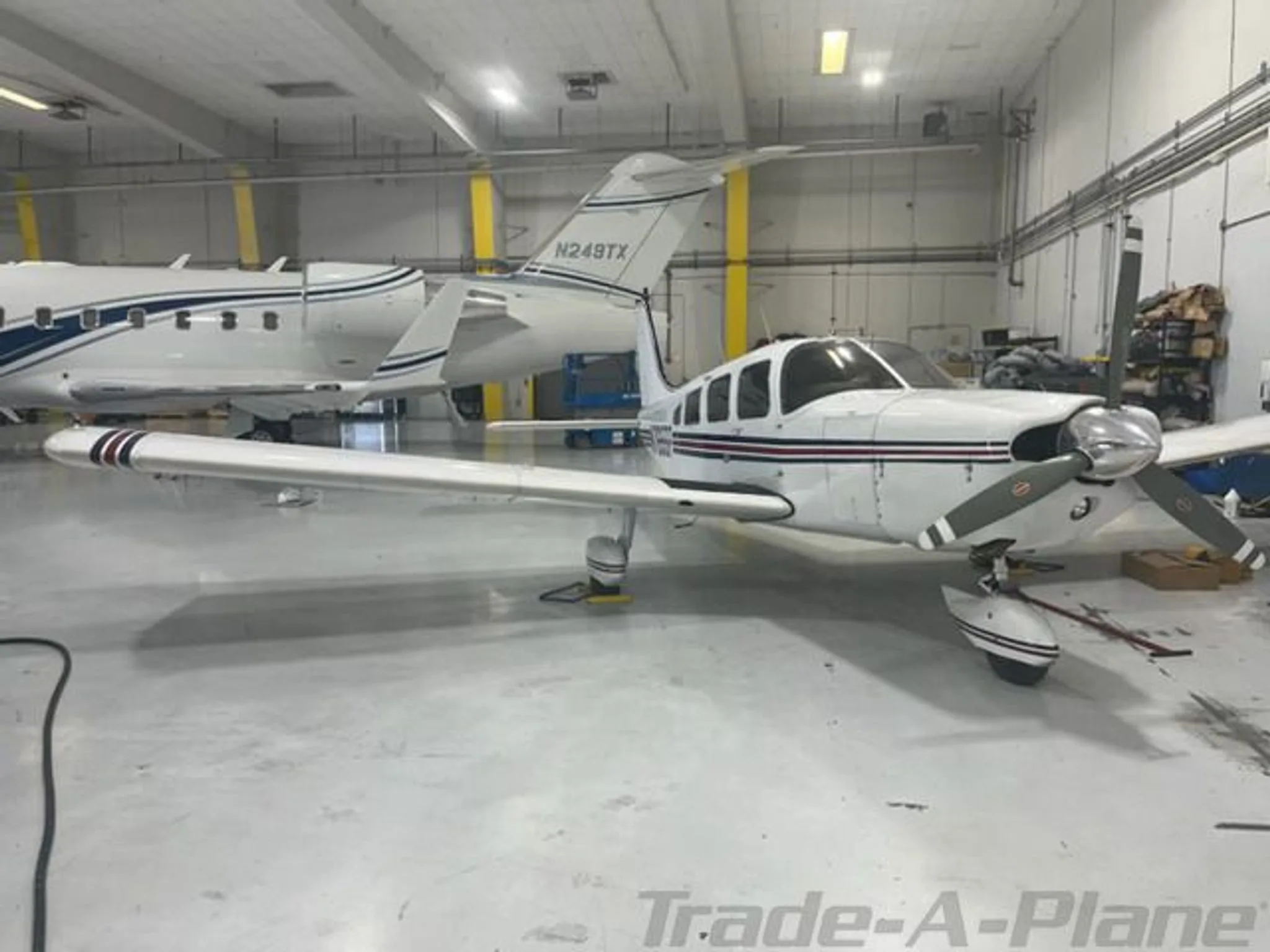 A white single-engine propeller aircraft with red and black stripes is parked in a hangar, with another jet aircraft in the background.