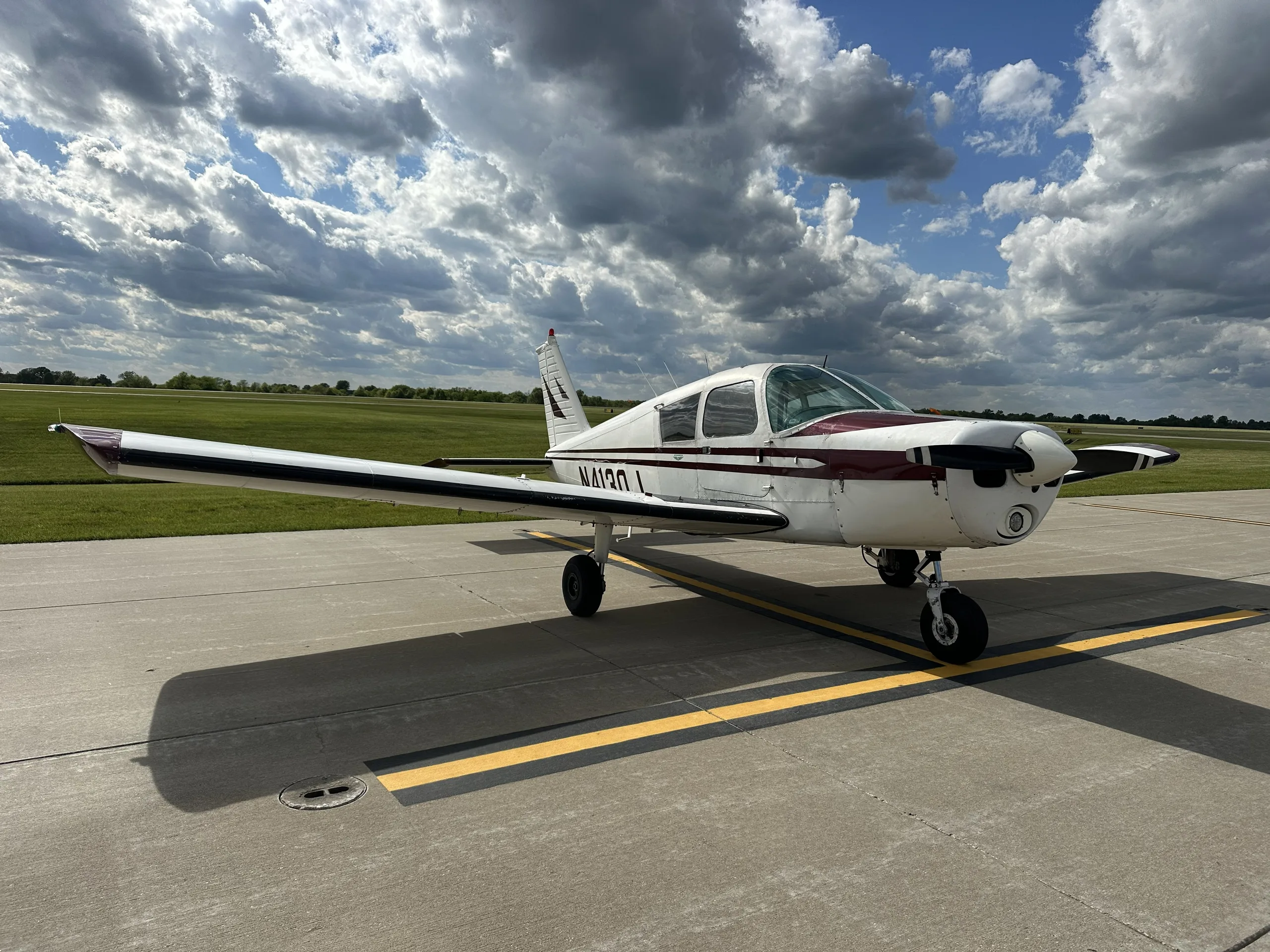 A white and maroon single-engine aircraft, likely a Piper Cherokee, parked on an asphalt ramp under a partly cloudy sky.