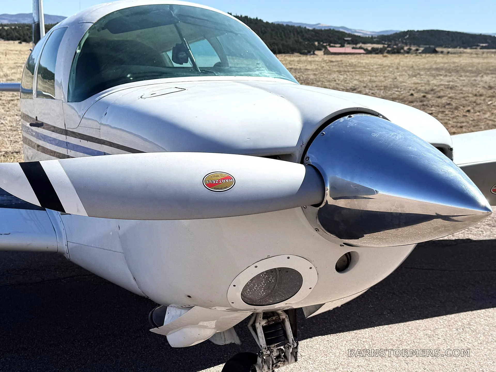 A close-up view of the front of a white Mooney M20J aircraft, showing the propeller, spinner, and landing gear.