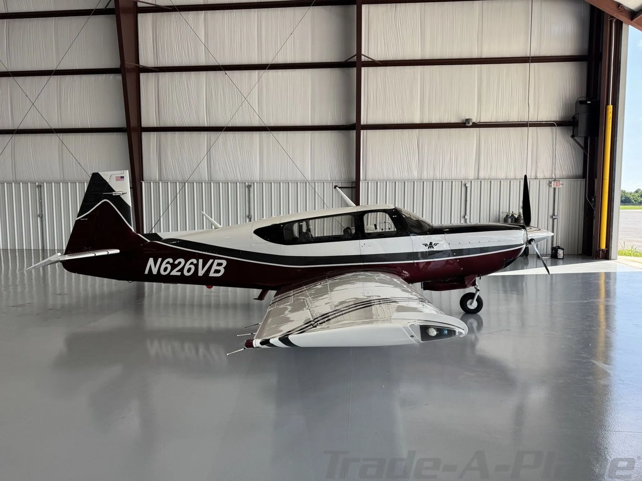 A Mooney aircraft with registration N626VB, featuring a maroon, white, and black paint scheme, is parked in a hangar with the door open to an airfield, viewed from the side.