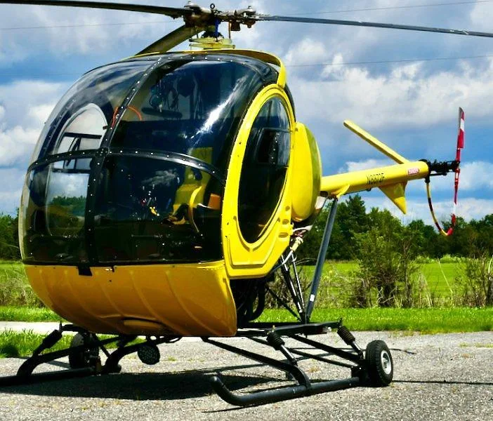 A bright yellow single-engine helicopter, N8872F, parked on an asphalt surface with a cloudy sky and green fields in the background.