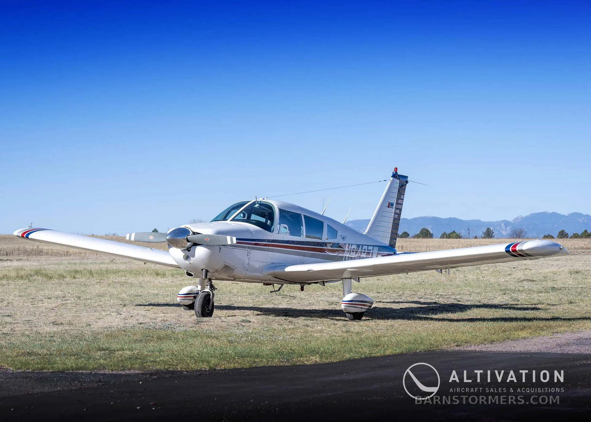A white single-engine propeller aircraft with blue and red stripes is parked on a grassy field with mountains in the background.