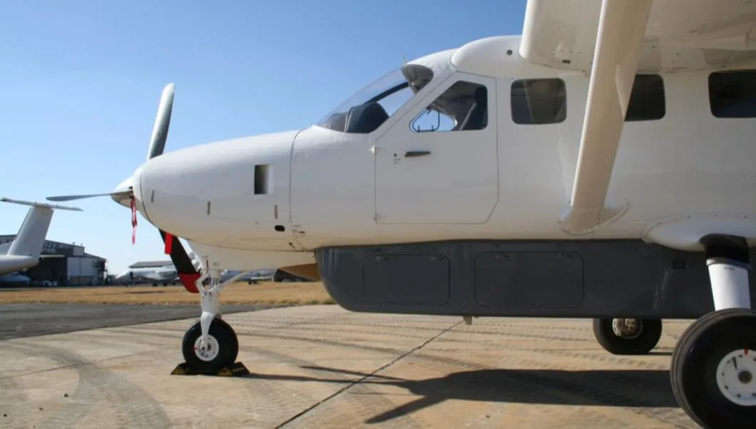 A white single-engine turboprop aircraft with a grey cargo pod is parked on an airport tarmac, viewed from the front-right side.