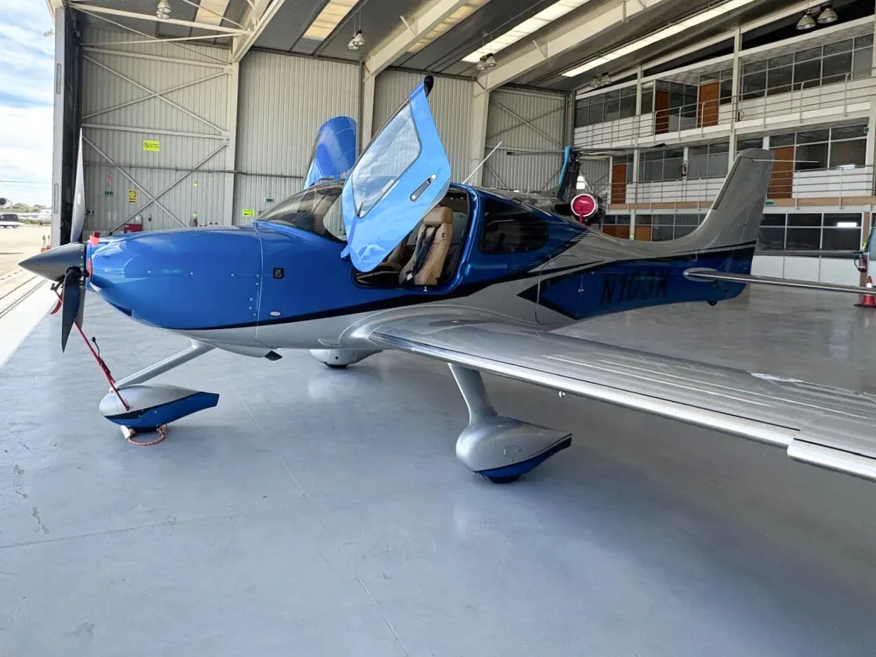 A blue and silver Cirrus SR22 aircraft (N103K) with open doors, viewed from the front-right in a hangar.