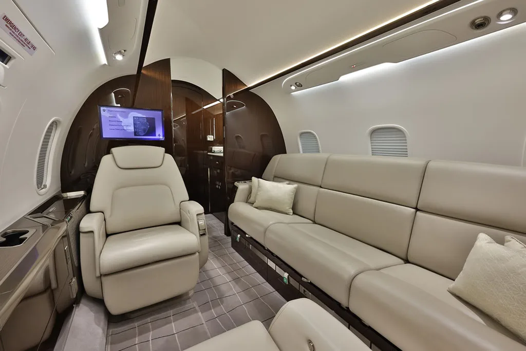 The private jet's cabin featuring a long beige leather sofa, a single armchair, and a wall-mounted screen.