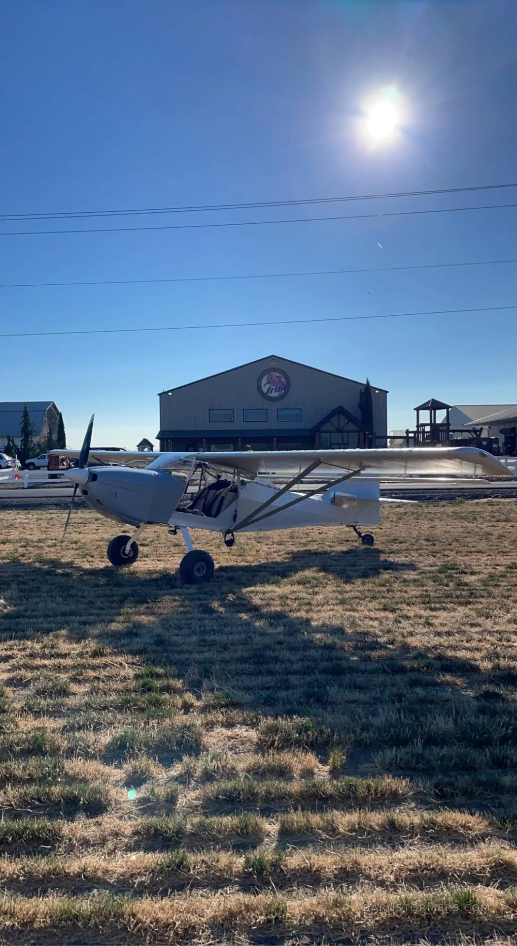 A light grey single-engine aircraft with a high wing and large tires is parked on dry grass in front of a large building under a bright blue sky.
