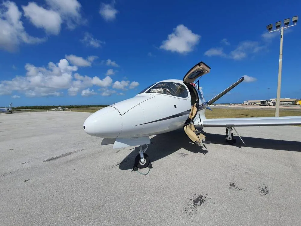 A white Cirrus Vision Jet with its main entry door open, parked on a concrete tarmac under a blue sky.