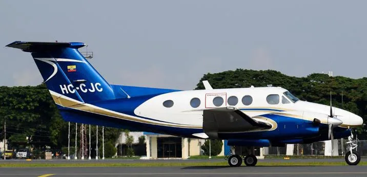 A blue and white Beechcraft King Air HC-CJC parked on a tarmac with buildings and trees.