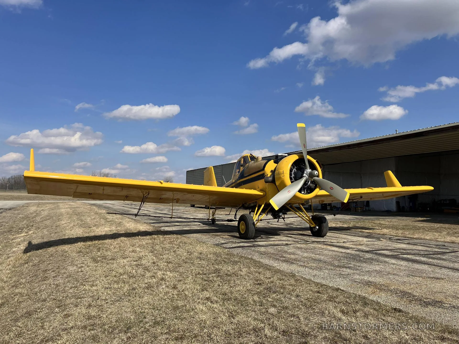 A yellow agricultural aircraft with a black stripe is parked on an asphalt ramp next to a hangar under a blue sky.