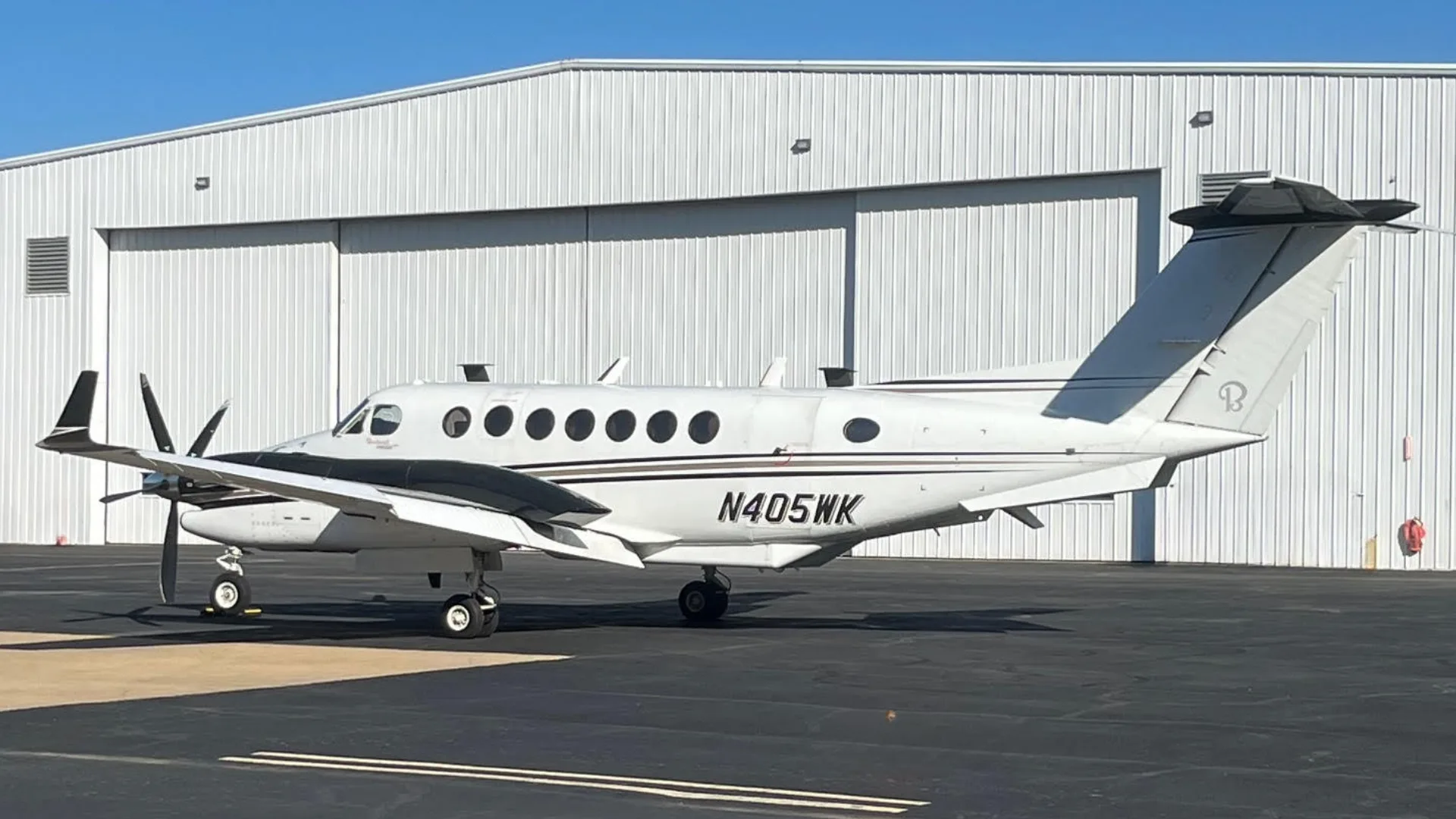 A side view of a white and black Beechcraft King Air N405WK parked on an asphalt tarmac in front of a white hangar.