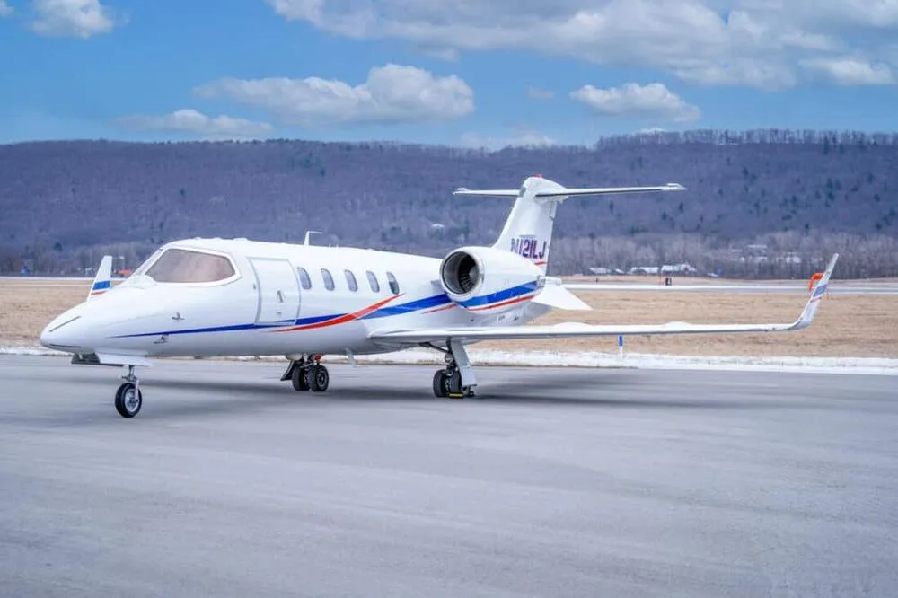 A white Learjet 45 with blue and red stripes on the fuselage and registration N121LJ, parked on a tarmac with mountains in the background under a blue sky.