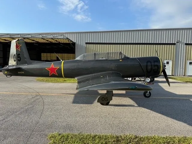A dark green military-style trainer aircraft with red star markings and '05' on the tail and cowling, parked on an asphalt ramp in front of hangars.