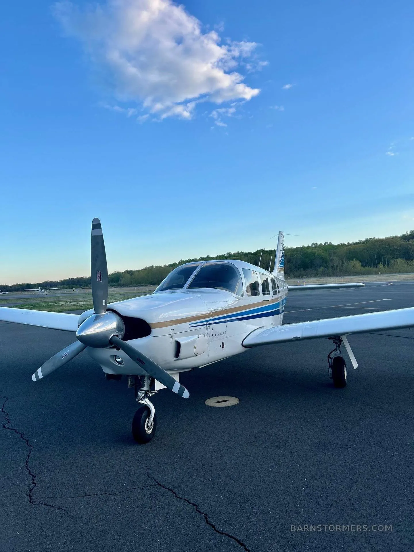 A white single-engine propeller aircraft with blue and gold stripes, viewed from the front-right, on an asphalt tarmac under a blue sky with clouds.