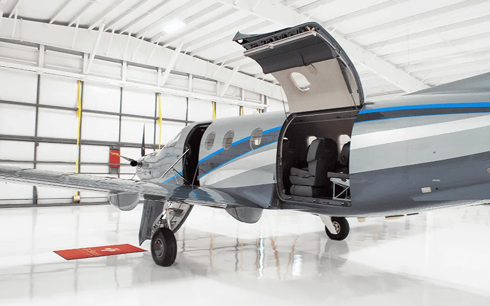 A silver Pilatus PC-12 turboprop aircraft with its passenger and cargo doors open, parked inside a white hangar.