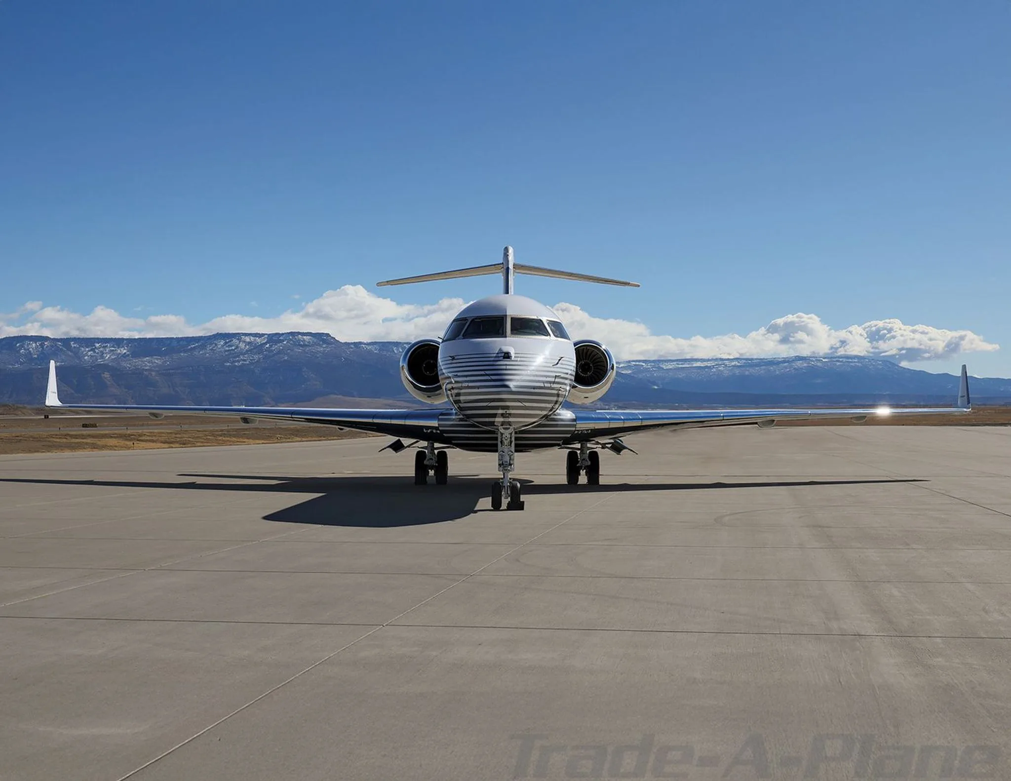 A head-on view of a white Bombardier Global 6000 business jet with grey pinstripes, parked on a tarmac with mountains in the background.