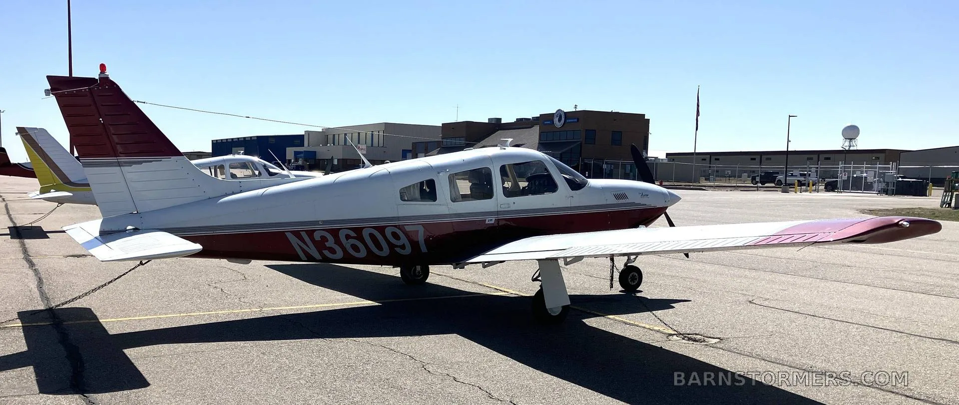 A side view of a white and maroon Piper Arrow aircraft (N36097) parked on an airport tarmac with buildings in the background.