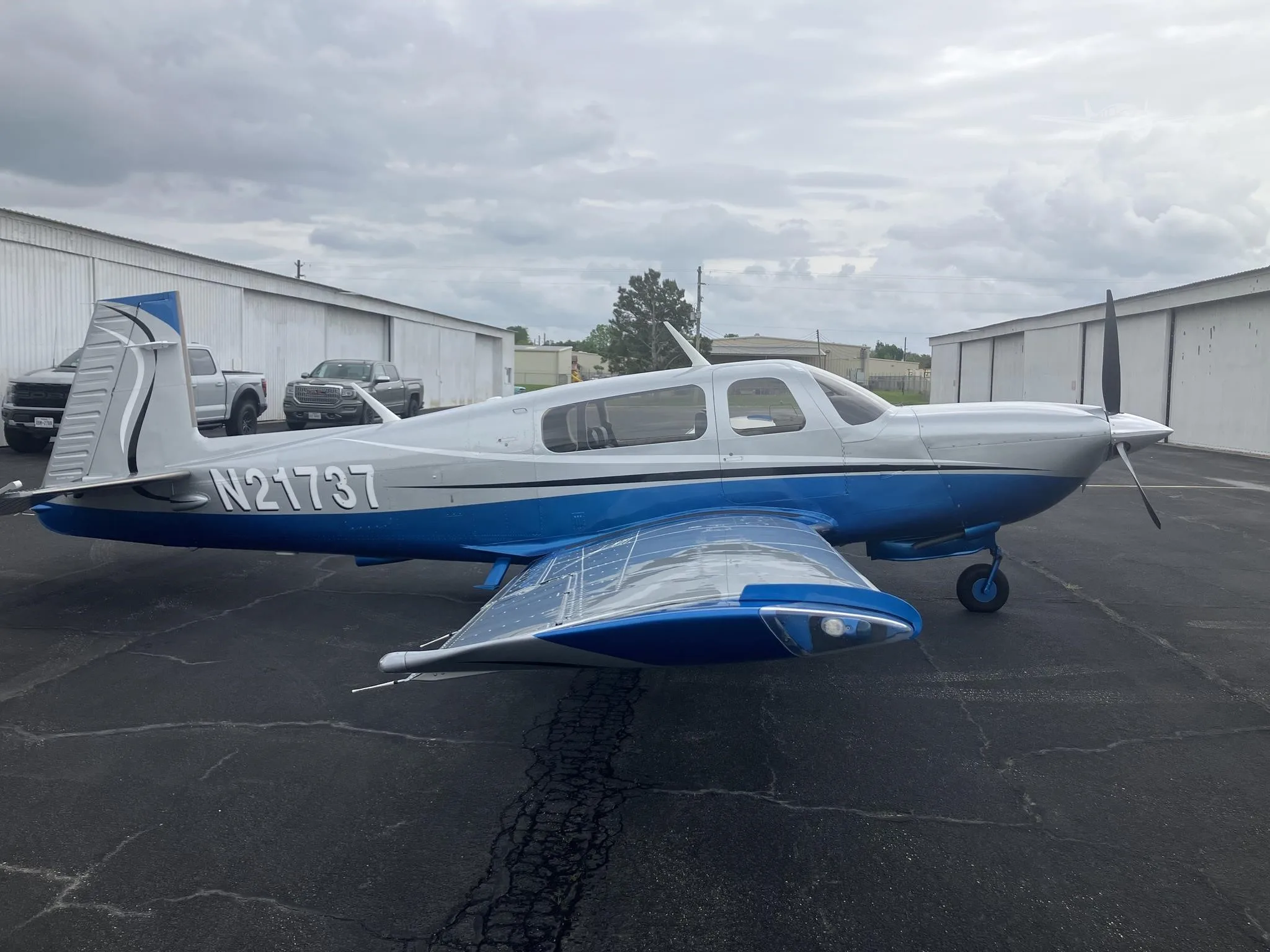 A silver and blue single-engine propeller aircraft, registration N21737, parked on an asphalt apron with hangars in the background under a cloudy sky.