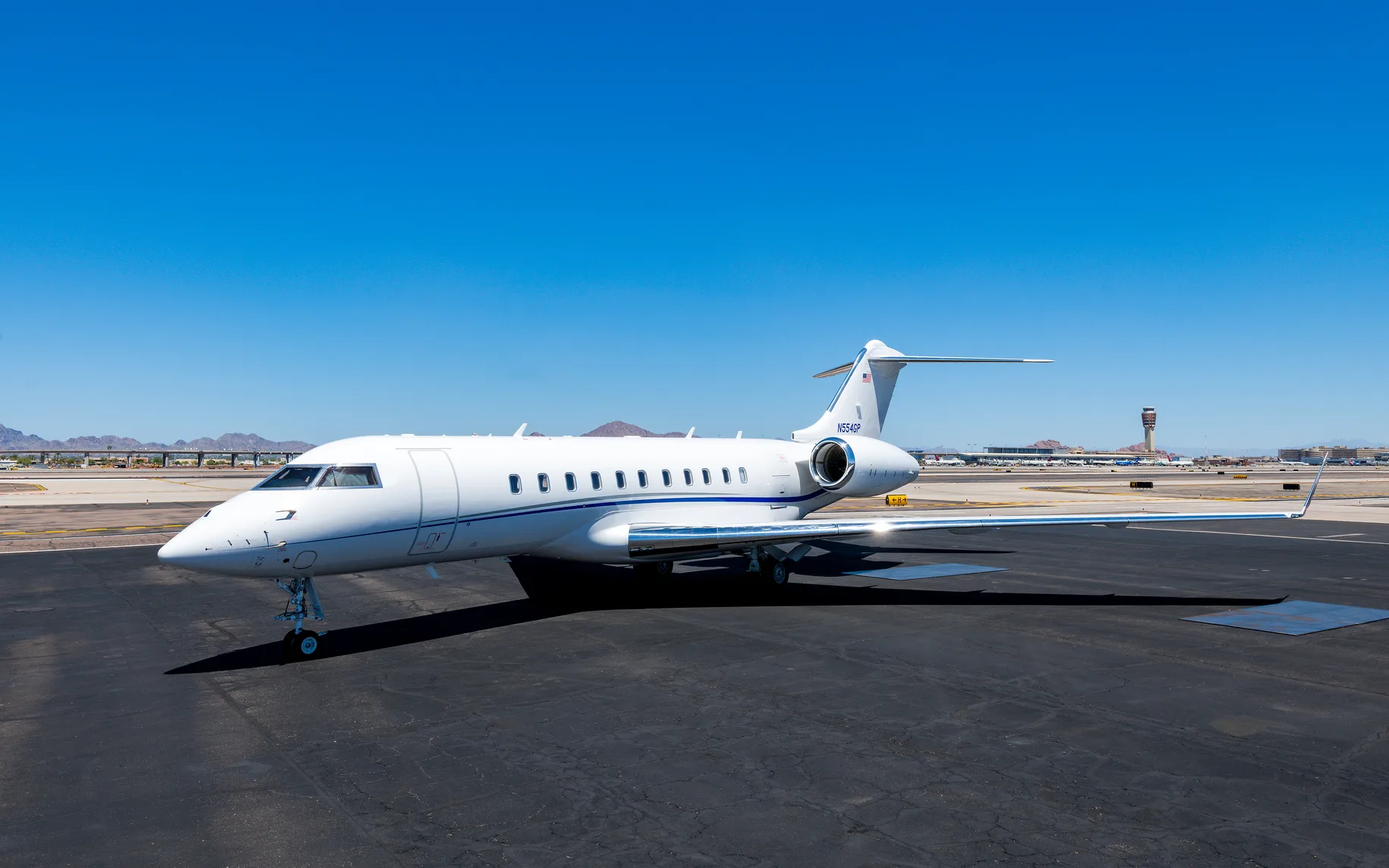 A white private jet with a blue stripe is parked on an airport tarmac on a sunny day, with mountains and a control tower in the background.