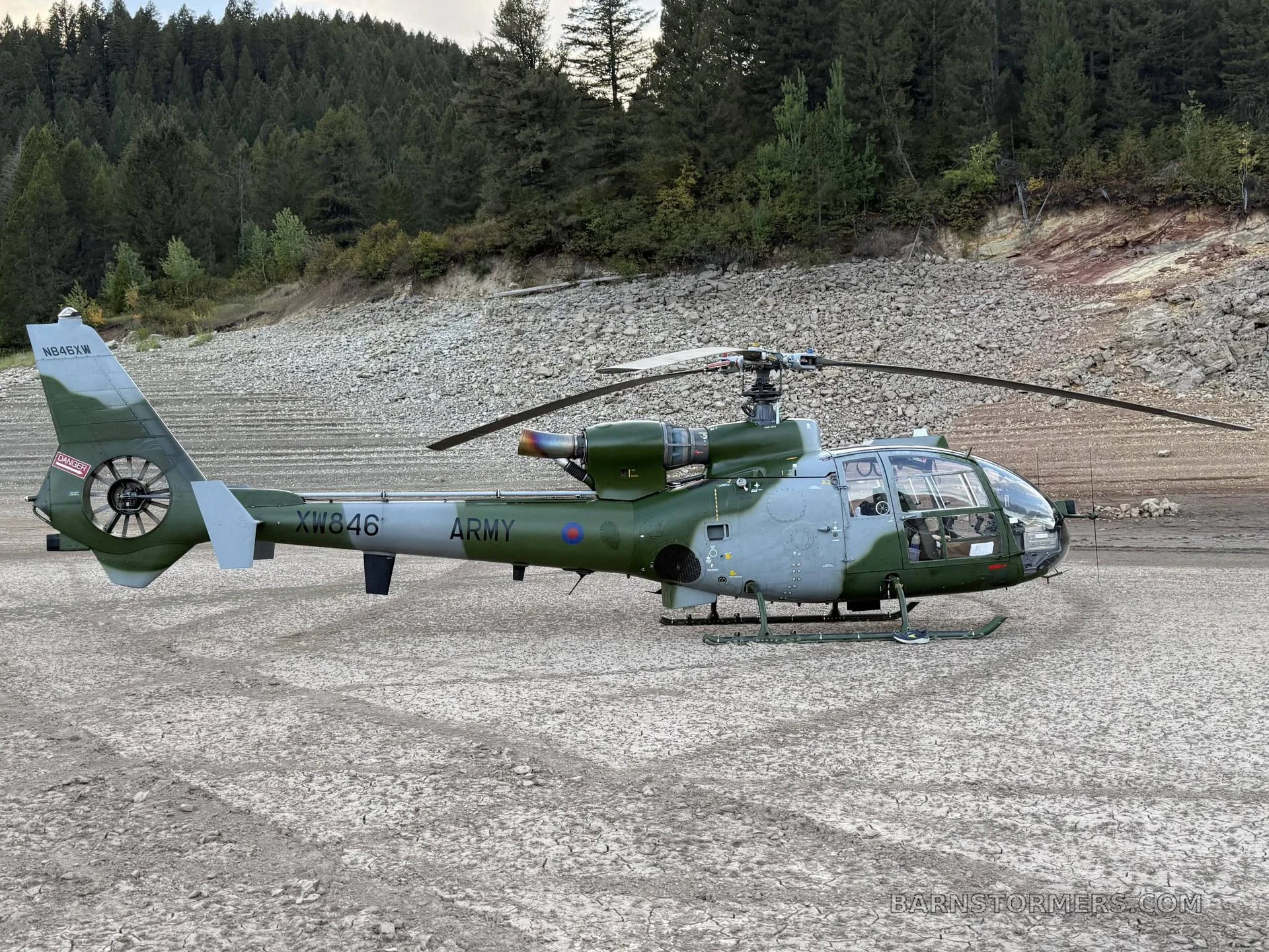 A green and grey military Westland Gazelle helicopter is parked on a cracked, dry ground next to a forest.